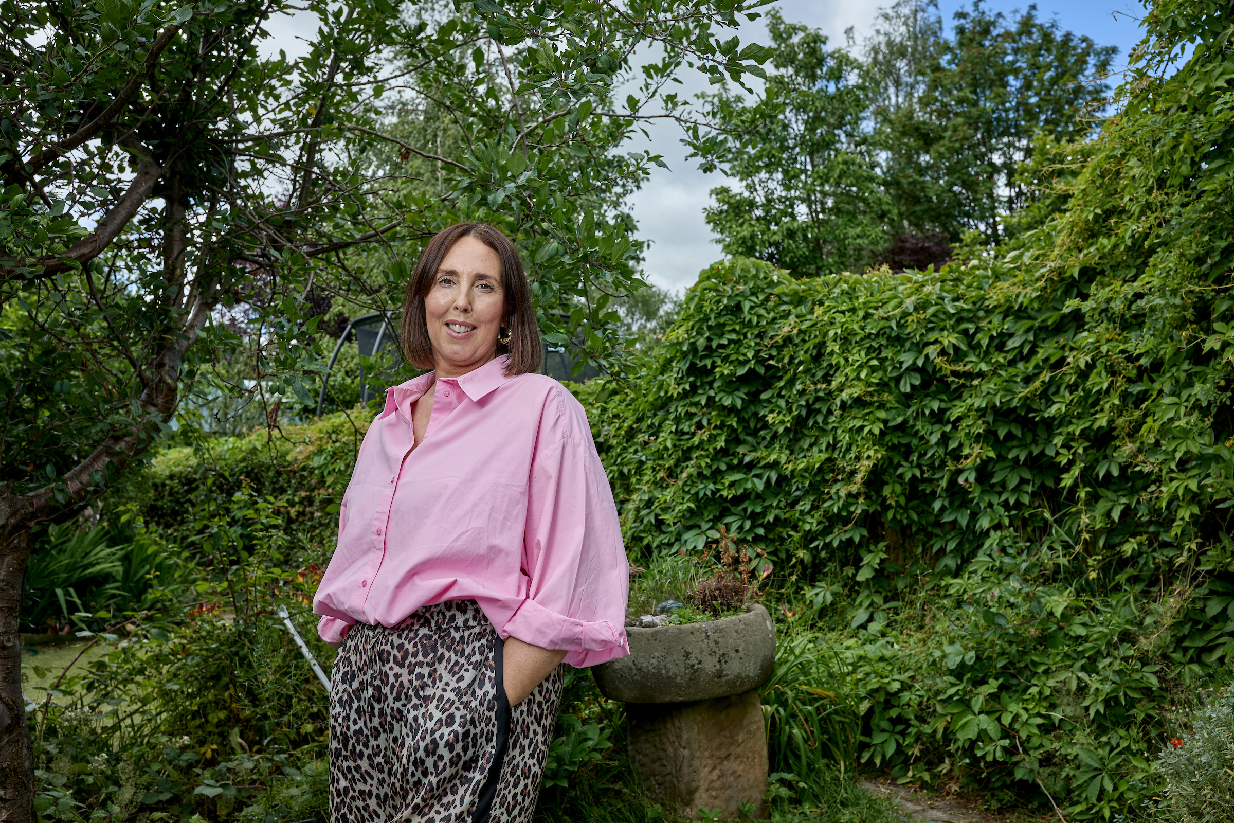 250629_icr_sally_steadman-south_0121 Sally stands in her garden surrounded by greenery. She smiles at the camera and wears a pink top