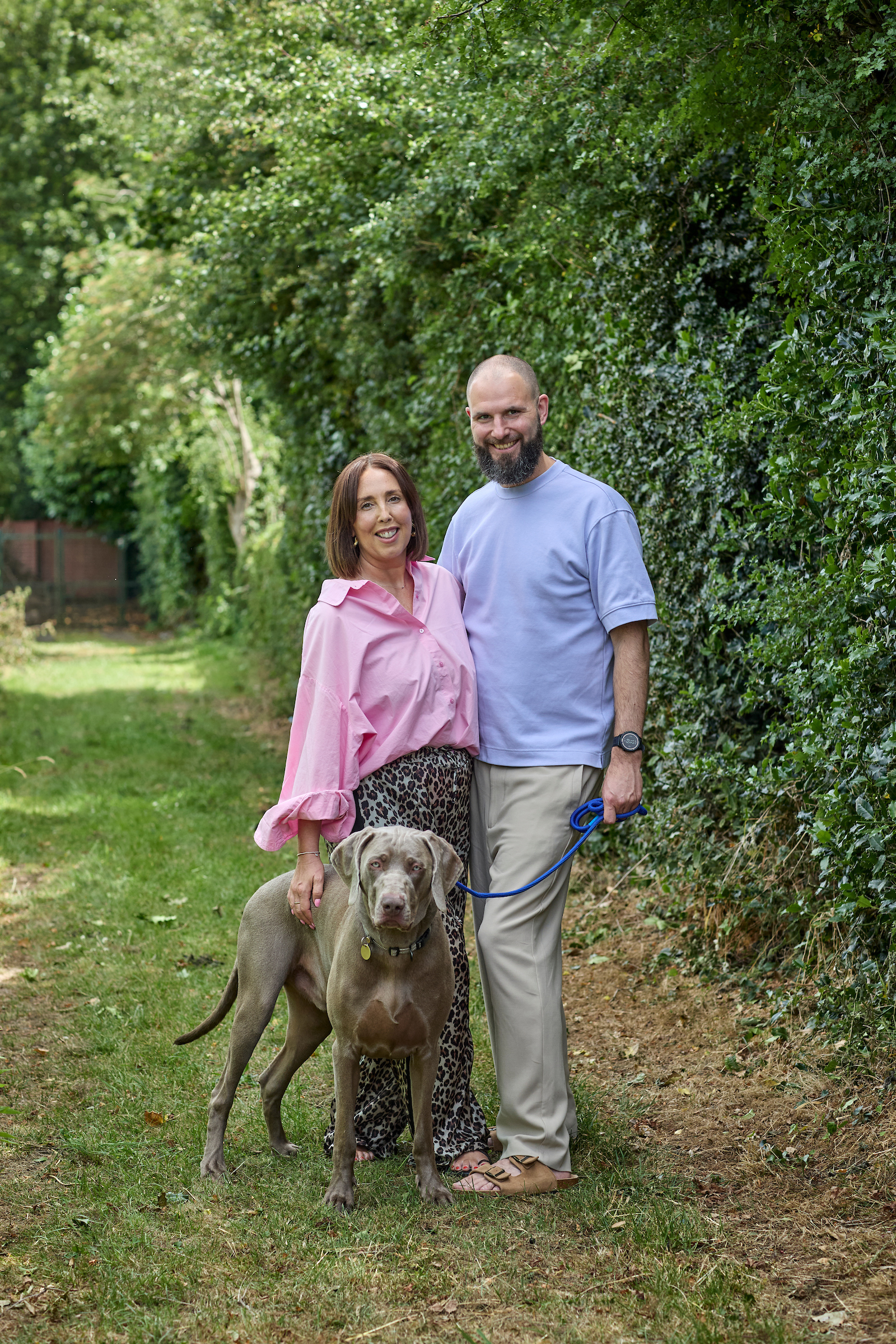 Sally and her husband stand in their garden with their dog, a grey Weimaraner