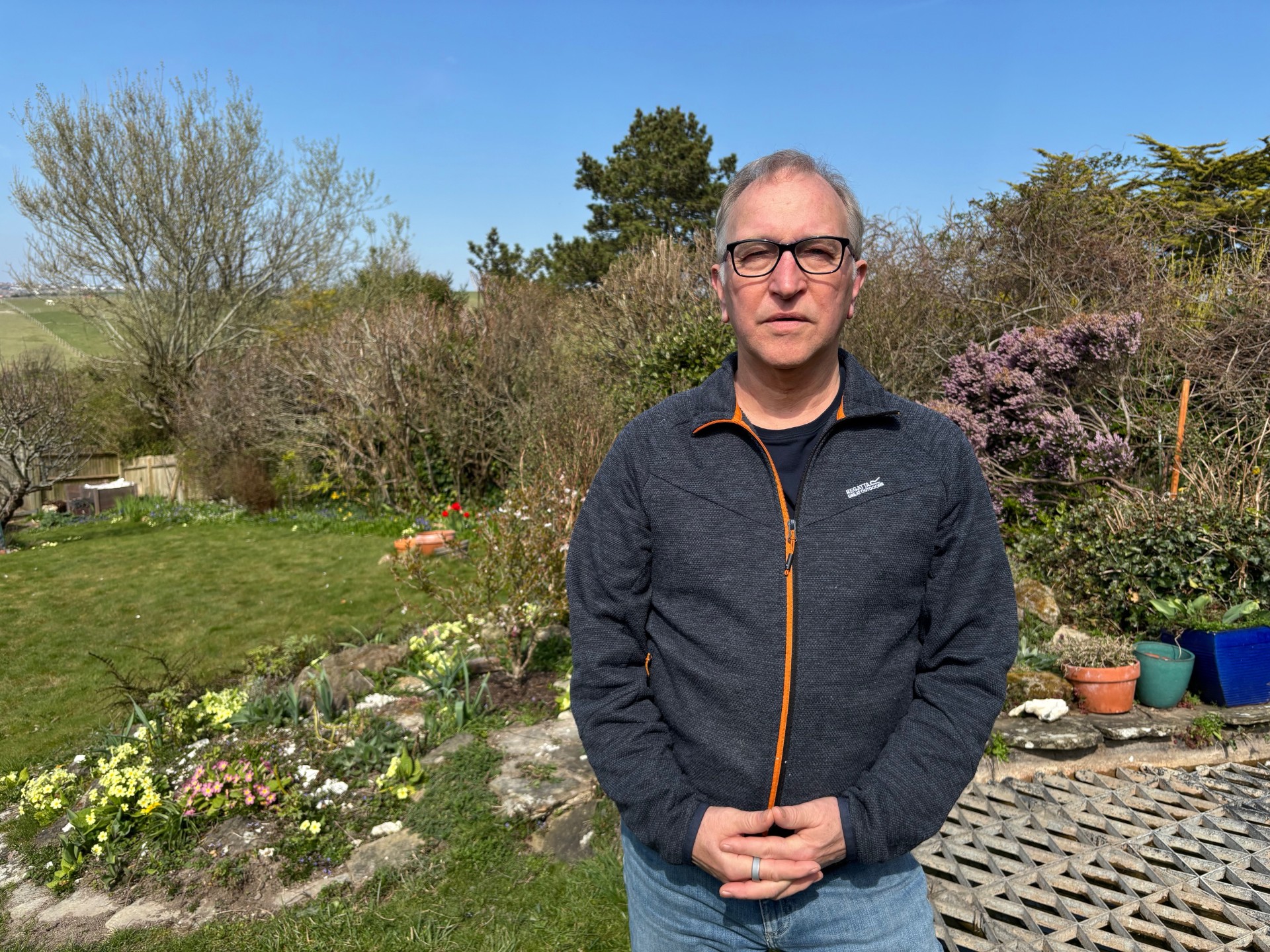 Giles Turner is wearing glasses, with his hands clasped in front of his body. He is in a garden.