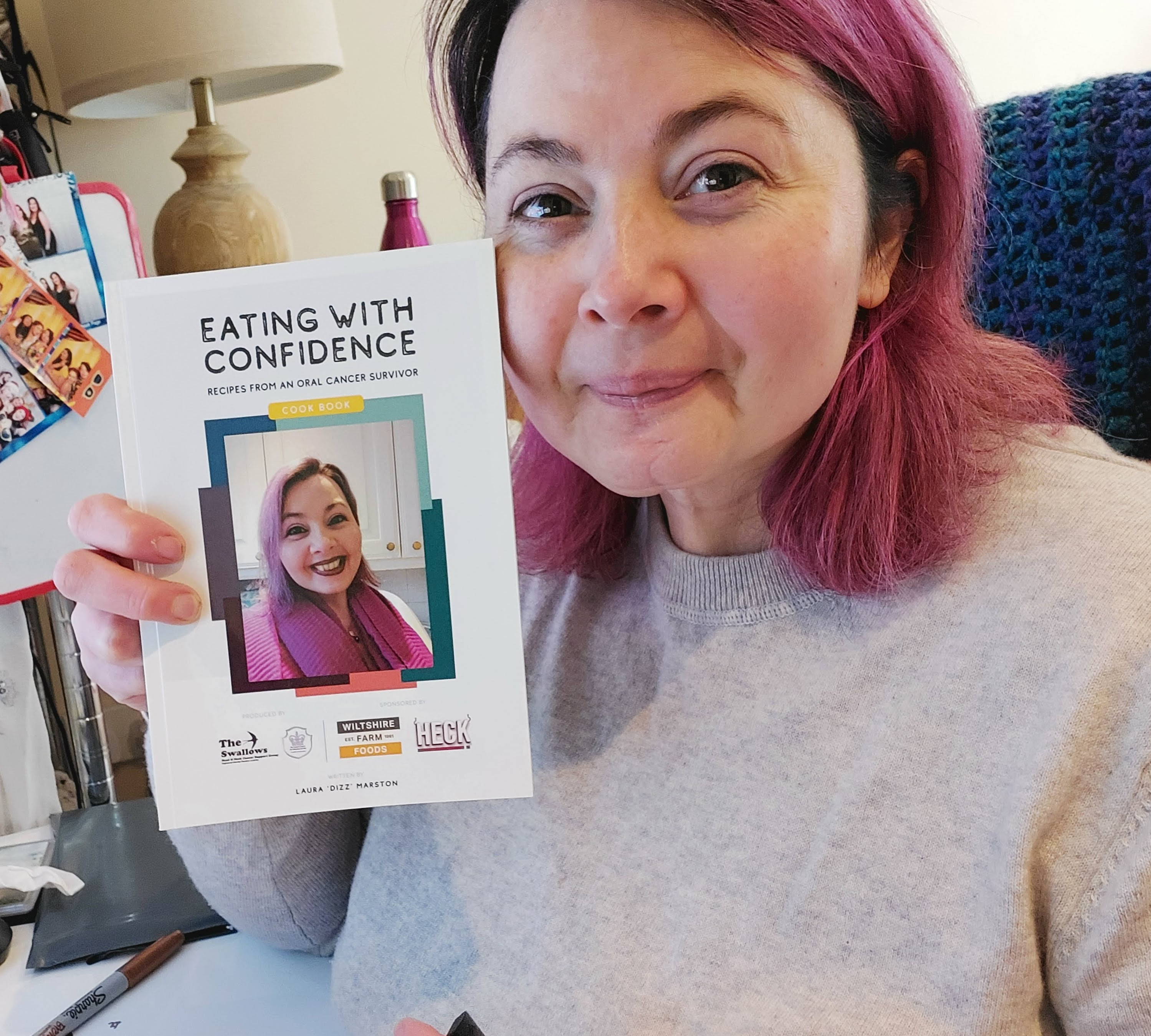 Laura holds up a piece up of paper which has a photo of her on it and says 'Eating with confidence, recipes from an oral cancer survivor'