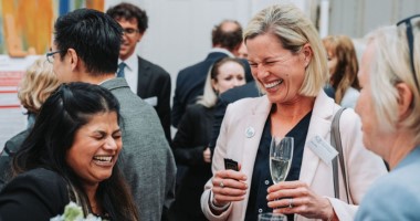 Summer reception 380x200px Three woman stand talking and laughing in a busy room. One holds a glass of champagne.
